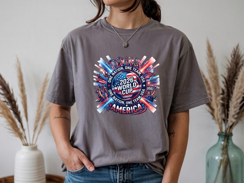 May include: A gray t-shirt featuring a colorful graphic for the 2026 World Cup in America. The design includes the text "2026 World Cup America" with red, white, and blue accents, and the words "One Nation, One Team, Goal!"