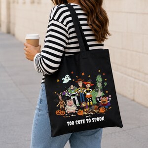 May include: A black tote bag featuring a colorful graphic of various cartoon characters dressed in Halloween costumes, including a ghost, pumpkins, and candy. The text "TOO CUTE TO SPOOK" is printed in white at the bottom of the bag design.