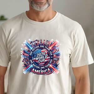 May include: Cream-colored t-shirt featuring a colorful graphic for the 2026 World Cup. The design includes a soccer ball, the text "2026 World Cup America", and the phrases "One Nation, One Team, USA" in red, white, and blue.