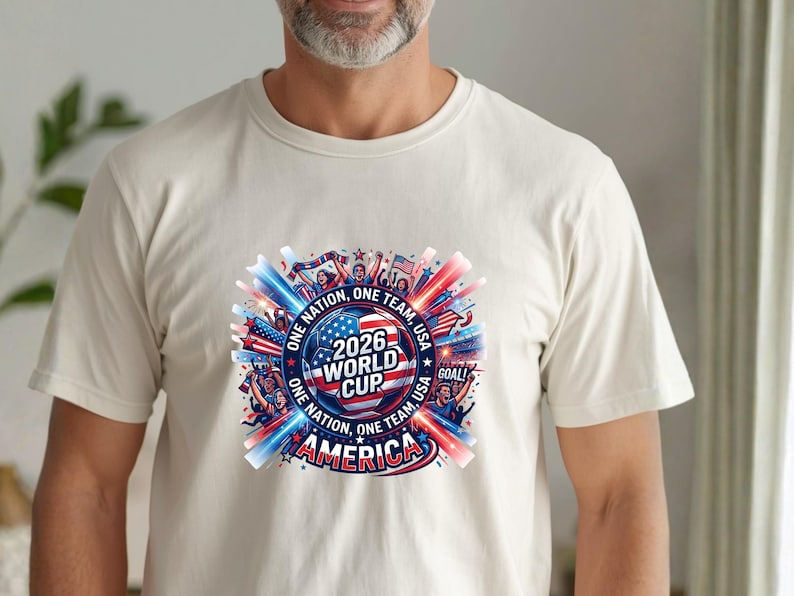 May include: Cream-colored t-shirt featuring a colorful graphic for the 2026 World Cup. The design includes a soccer ball, the text "2026 World Cup America", and the phrases "One Nation, One Team, USA" in red, white, and blue.