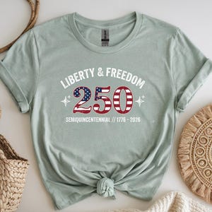 Puede incluir: Camiseta verde claro con el texto "Liberty & Freedom 250" en blanco y rojo, con un dise&ntilde;o que recuerda a la bandera estadounidense. La camiseta tambi&eacute;n incluye el texto "Semiquincentennial // 1776-2026".