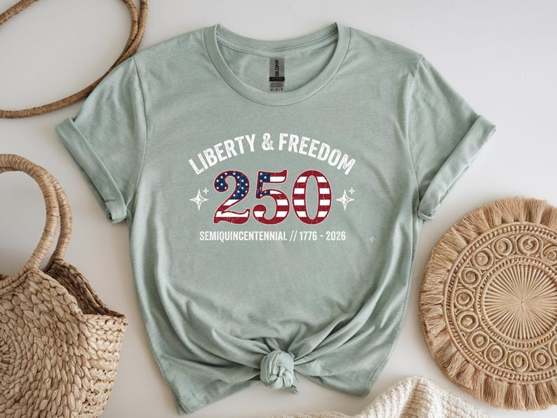Puede incluir: Camiseta verde claro con el texto "Liberty & Freedom 250" en blanco y rojo, con un dise&ntilde;o que recuerda a la bandera estadounidense. La camiseta tambi&eacute;n incluye el texto "Semiquincentennial // 1776-2026".