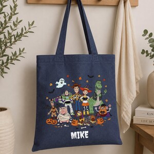May include: A dark blue canvas tote bag featuring a colorful graphic of Toy Story characters dressed in Halloween costumes, including pumpkins, a ghost, and bats. The name MIKE is printed in white capital letters at the bottom of the design.