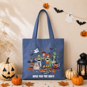 May include: A blue canvas tote bag featuring a colorful graphic of Toy Story characters dressed in Halloween costumes, including Woody, Buzz Lightyear, and Jessie. The text "HERE FOR THE BOO'S" is printed in white below the character illustration.