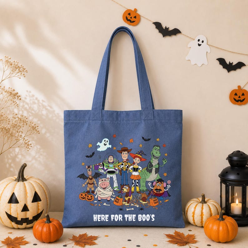 May include: A blue canvas tote bag featuring a colorful graphic of Toy Story characters dressed in Halloween costumes, including Woody, Buzz Lightyear, and Jessie. The text "HERE FOR THE BOO'S" is printed in white below the character illustration.