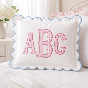 May include: A rectangular white throw pillow featuring a scalloped edge trimmed in light blue piping. The center of the pillow displays the letters A, B, and C embroidered in a pink and white gingham pattern with a decorative serif font style.