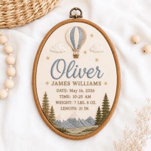 May include: An oval embroidery hoop featuring a cream fabric background with a blue and white hot air balloon, clouds, and stars. The text reads: Oliver, JAMES WILLIAMS, DATE: May 16, 2026, TIME: 10:25 AM, WEIGHT: 7 LBS. 8 OZ., LENGTH: 21 IN. with pine trees and mountains.