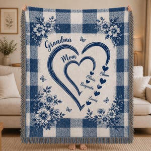 May include: A blue and white plaid throw blanket with floral accents and the words "Grandma Mom" and names inside a heart design. The blanket has a fringe edge and is held up by a person.