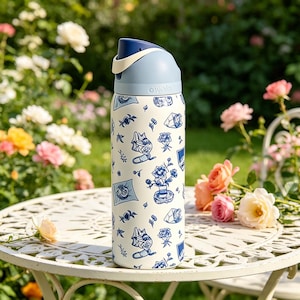 Op de afbeelding: Een wit en blauw ge&iuml;soleerde waterfles met een bloemen- en kattenontwerp. De fles heeft een blauwe dop en een cr&egrave;mekleurige handgreep. De fles staat op een witte metalen tafel met rozen op de achtergrond.