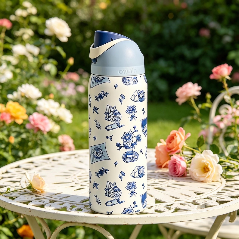 Op de afbeelding: Een wit en blauw ge&iuml;soleerde waterfles met een bloemen- en kattenontwerp. De fles heeft een blauwe dop en een cr&egrave;mekleurige handgreep. De fles staat op een witte metalen tafel met rozen op de achtergrond.