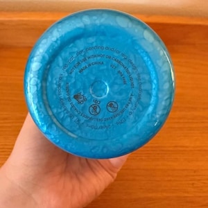 Op de afbeelding: De bodem van een blauwe waterfles met een textuur, bubbelachtig patroon. De onderkant van de fles heeft bedrukte tekst, waaronder "MADE IN CHINA", "BPA FREE" en waarschuwingen over hete of koolzuurhoudende vloeistoffen. De fles bevat 710 ml.
