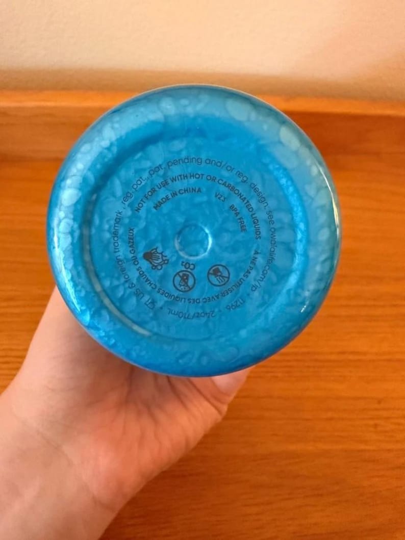 Op de afbeelding: De bodem van een blauwe waterfles met een textuur, bubbelachtig patroon. De onderkant van de fles heeft bedrukte tekst, waaronder "MADE IN CHINA", "BPA FREE" en waarschuwingen over hete of koolzuurhoudende vloeistoffen. De fles bevat 710 ml.
