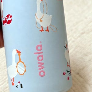 K&ouml;nnte beinhalten: Eine hellblaue Wasserflasche mit einem sich wiederholenden Muster aus wei&szlig;en G&auml;nsen mit orangefarbenen Schn&auml;beln und F&uuml;&szlig;en. Die G&auml;nse sind mit verschiedenen Accessoires wie einer Perlenkette, einem Handspiegel und rotem Lippenstift abgebildet. Das Wort owala ist in Pink gedruckt.