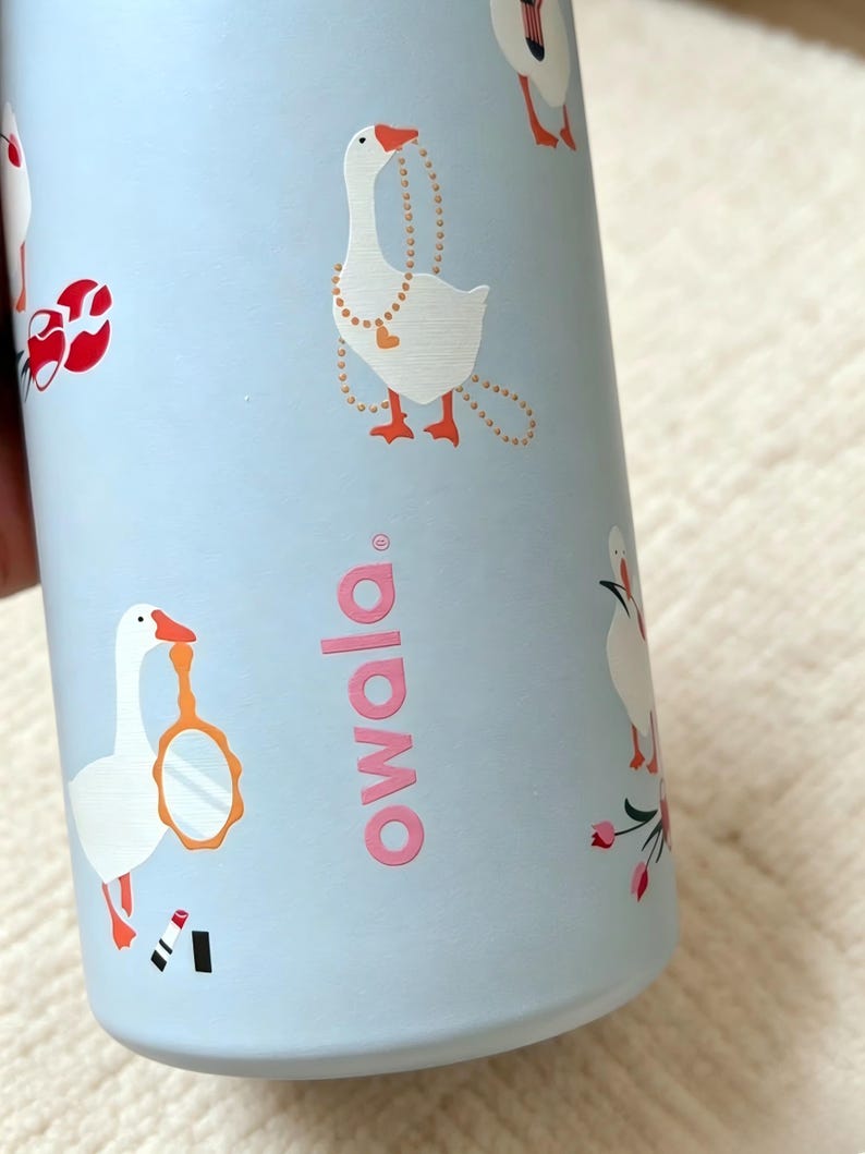 K&ouml;nnte beinhalten: Eine hellblaue Wasserflasche mit einem sich wiederholenden Muster aus wei&szlig;en G&auml;nsen mit orangefarbenen Schn&auml;beln und F&uuml;&szlig;en. Die G&auml;nse sind mit verschiedenen Accessoires wie einer Perlenkette, einem Handspiegel und rotem Lippenstift abgebildet. Das Wort owala ist in Pink gedruckt.
