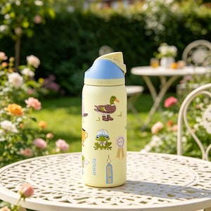 May include: A yellow Owala water bottle with a blue and yellow lid, decorated with cartoon animal stickers. The stickers include a duck, frog, and a coffee cup. The bottle is sitting on a white metal table in a garden setting.