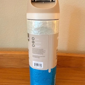 Op de afbeelding: Een herbruikbare waterfles met een beige deksel en bovenkant, een getextureerd parelmoer middenstuk en een getextureerde blauwe basis. De fles heeft een klapdeksel en een label met tekst en pictogrammen. BPA-vrij.