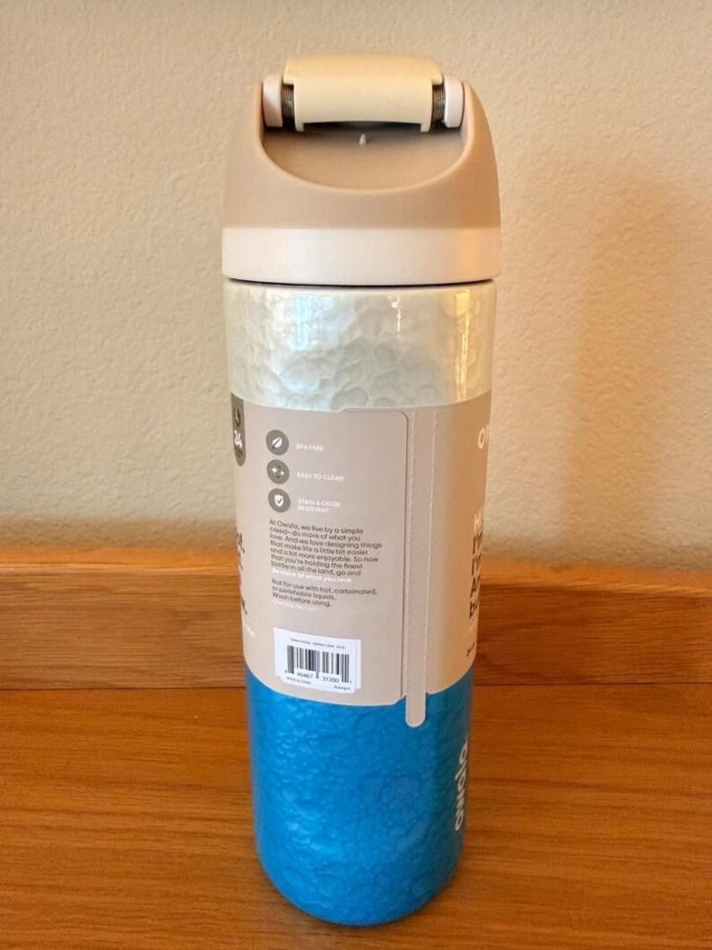 Op de afbeelding: Een herbruikbare waterfles met een beige deksel en bovenkant, een getextureerd parelmoer middenstuk en een getextureerde blauwe basis. De fles heeft een klapdeksel en een label met tekst en pictogrammen. BPA-vrij.