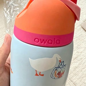 K&ouml;nnte beinhalten: Eine hellblaue Owala-Trinkflasche mit einem wiederkehrenden Muster aus wei&szlig;en G&auml;nsen. Eine Gans tr&auml;gt ein blaues Netz mit Lebensmitteln. Die Flasche hat einen leuchtend orangefarbenen und rosafarbenen Deckel mit Tragegriff und Markennamen.