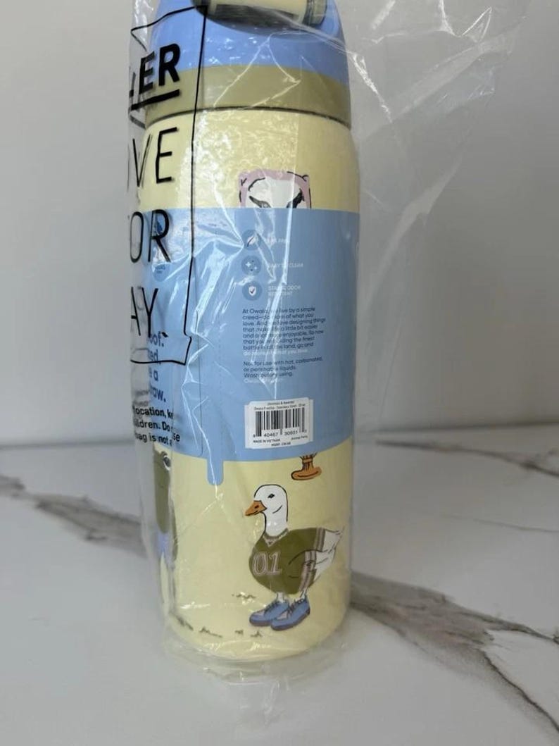 May include: A yellow and blue water bottle with a duck graphic wearing a jersey and sneakers. The bottle is wrapped in plastic and has the words "ER VE OR AY" visible. The bottle is designed for everyday use.