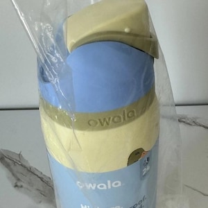 May include: A blue and yellow Owala insulated water bottle with a built-in straw. The bottle has a light blue body, a yellow lid, and a green accent band. The bottle is 32 oz. and leak proof.