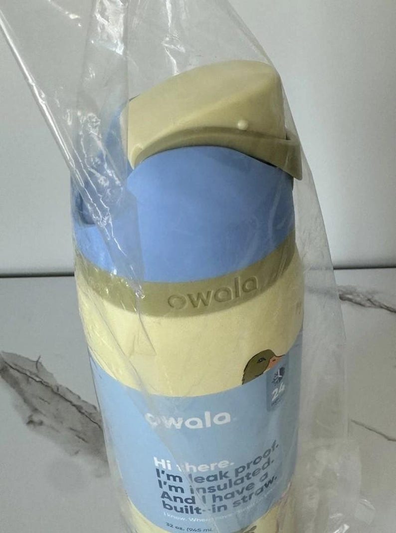 May include: A blue and yellow Owala insulated water bottle with a built-in straw. The bottle has a light blue body, a yellow lid, and a green accent band. The bottle is 32 oz. and leak proof.