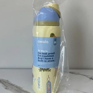 May include: A yellow and blue Owala water bottle with a built-in straw, packaged in clear plastic. The bottle has the text "Hi there. I'm leak proof. I'm insulated. And I have a built-in straw."