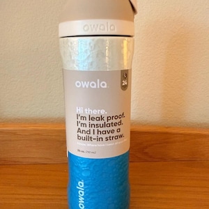 Op de afbeelding: Een blauwe, beige en witte Owala waterfles. De fles heeft een getextureerde blauwe basis, een zilveren middengedeelte en een beige bovenkant met een ingebouwd rietje. De tekst op de fles luidt: "Hallo. Ik ben lekvrij. Ik ben ge&iuml;soleerd. En ik heb een ingebouwd rietje."