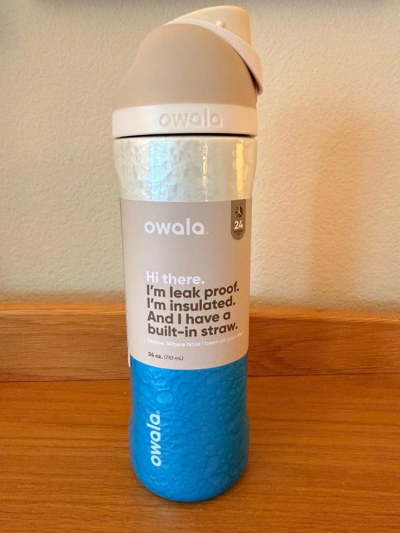 Op de afbeelding: Een blauwe, beige en witte Owala waterfles. De fles heeft een getextureerde blauwe basis, een zilveren middengedeelte en een beige bovenkant met een ingebouwd rietje. De tekst op de fles luidt: "Hallo. Ik ben lekvrij. Ik ben ge&iuml;soleerd. En ik heb een ingebouwd rietje."