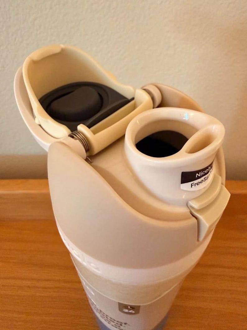 Op de afbeelding: Een beige reismok met een open deksel, waarbinnen een donker interieur zichtbaar is. Het deksel heeft een scharnierend ontwerp met een zwarte rubberen afdichting. De mok heeft een kleine tuit en een label met de tekst "Nicely FreeS".