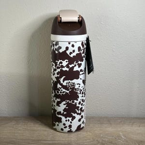 May include: A stainless steel travel mug with a brown and white cow print design. The lid is brown and beige, with a flip-top opening. A black tag hangs from the side. The mug is cylindrical and stands on a wooden surface.