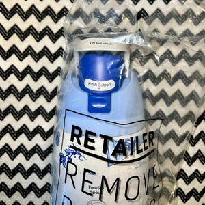 Op de afbeelding: Een blauw-witte waterfles met een drukknop en de tekst "Lift to Unlock". De fles zit in een doorzichtige plastic zak met de tekst "RETAILER REMOVE".