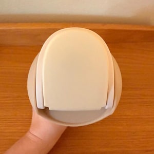 Op de afbeelding: Een cr&egrave;mekleurige, afgeronde container met een platte bovenkant en een gebogen basis. De container heeft aan elke kant een witte handgreep. Het object wordt door een persoon vastgehouden.
