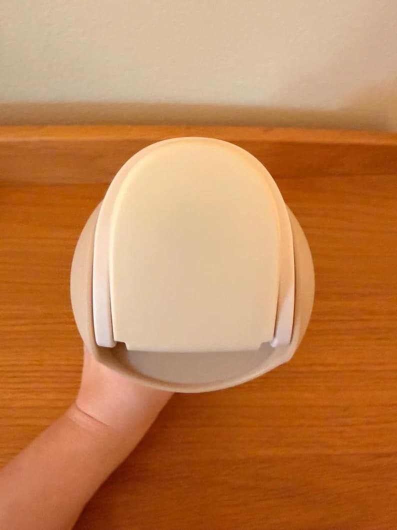 Op de afbeelding: Een cr&egrave;mekleurige, afgeronde container met een platte bovenkant en een gebogen basis. De container heeft aan elke kant een witte handgreep. Het object wordt door een persoon vastgehouden.