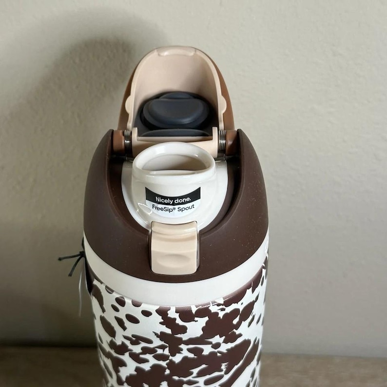 May include: A brown and white patterned water bottle with a flip-top lid. The bottle has a brown and white cow print design on the lower half. The lid is brown and tan with a black spout. The text "Nicely done. FreeSip Spout" is visible.