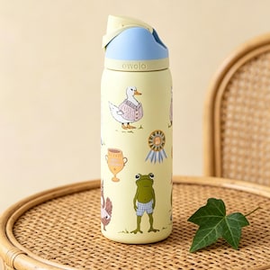 May include: A yellow Owala water bottle with a blue and light yellow lid. The bottle is decorated with cartoon animals, including a duck in a sweater and a frog in blue shorts. The bottle is on a wicker table.