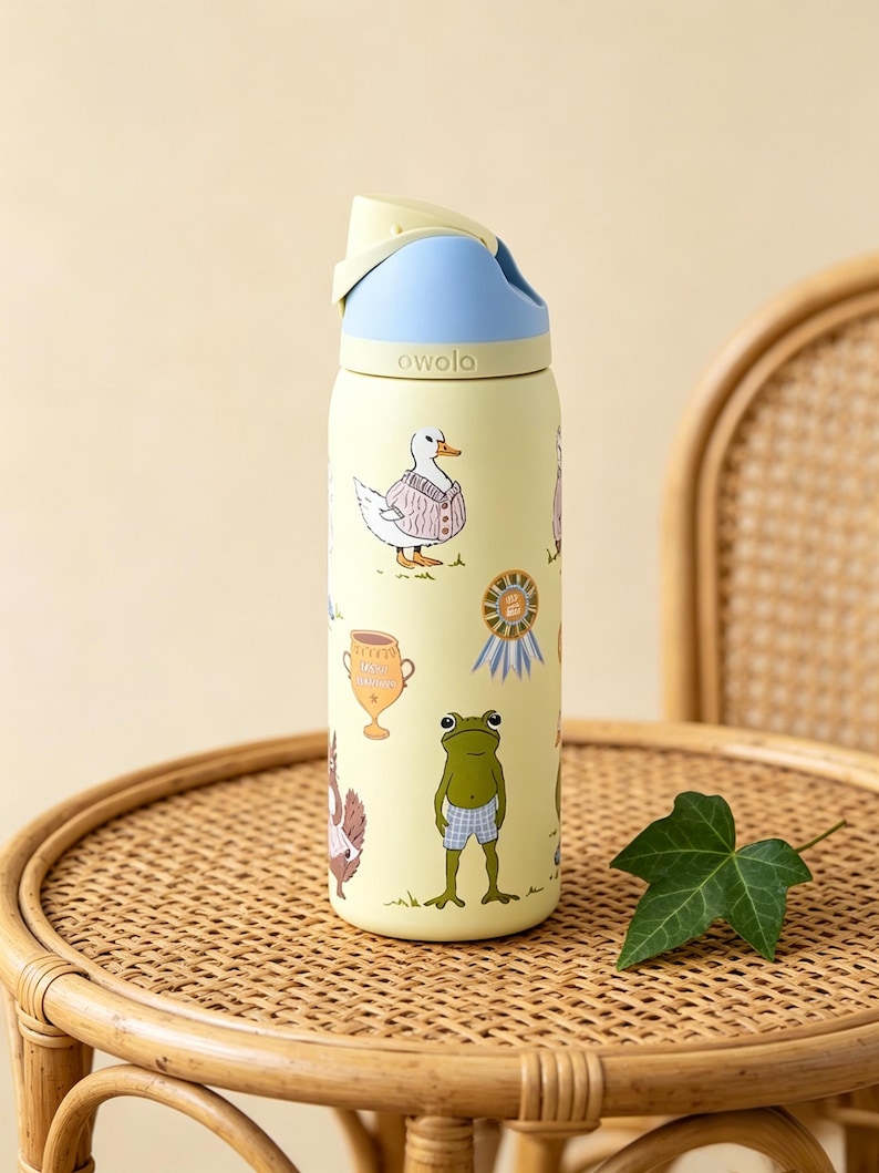 May include: A yellow Owala water bottle with a blue and light yellow lid. The bottle is decorated with cartoon animals, including a duck in a sweater and a frog in blue shorts. The bottle is on a wicker table.