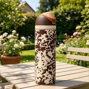 May include: A stainless steel Owala water bottle with a brown and cream cow print design. The bottle has a brown top with a tan handle and a white band. The bottle is sitting on a wooden table in a garden setting.