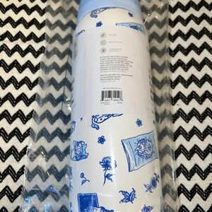 Op de afbeelding: Witte waterfles met een lichtblauw deksel, versierd met blauwe bloemen- en kattenillustraties. De fles is verpakt in doorzichtig plastic. De fles heeft tekst, inclusief de merknaam en productinformatie.