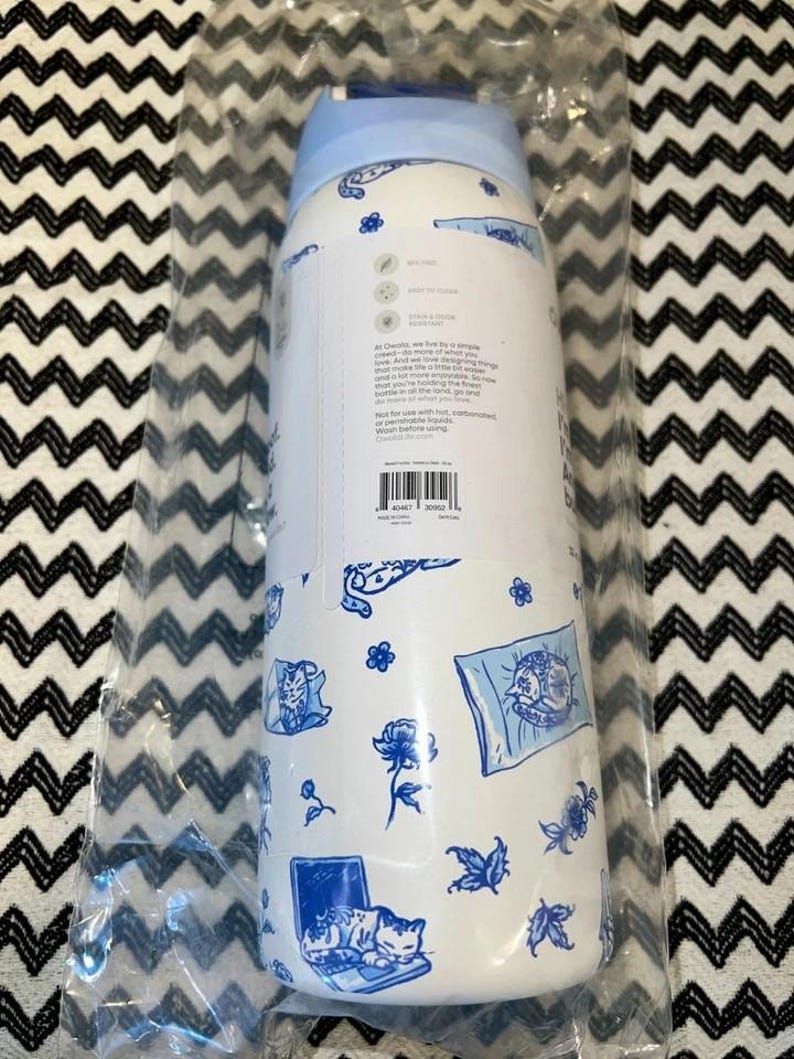 Op de afbeelding: Witte waterfles met een lichtblauw deksel, versierd met blauwe bloemen- en kattenillustraties. De fles is verpakt in doorzichtig plastic. De fles heeft tekst, inclusief de merknaam en productinformatie.