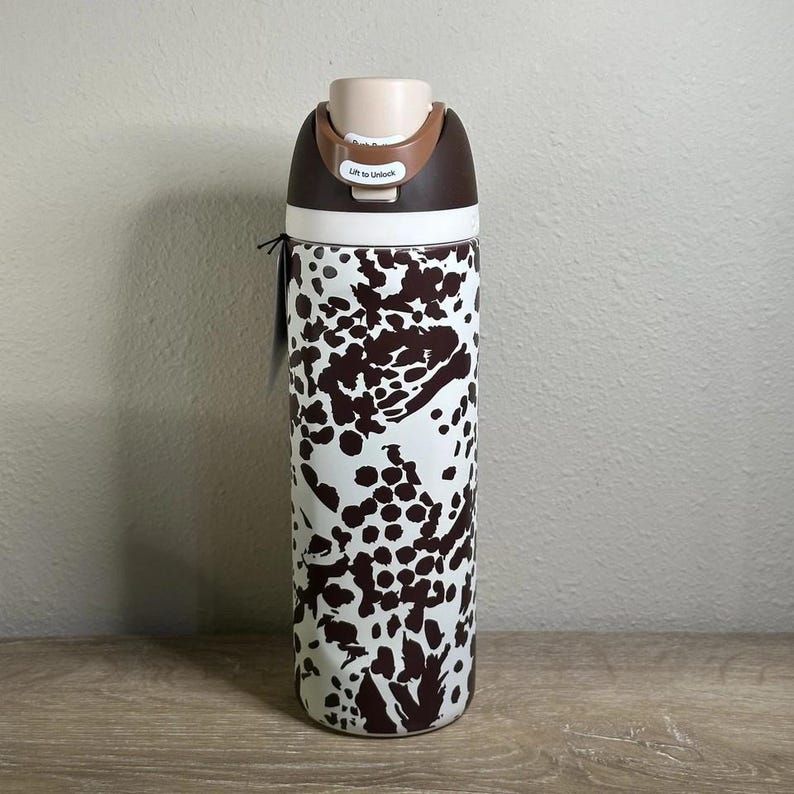 May include: A stainless steel water bottle with a white and brown cow print design. The lid is brown and beige with the text "Lift to Unlock". The bottle is cylindrical and has a black tag attached.