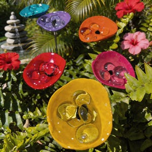 Könnte beinhalten: Eine Sammlung farbenfroher Gartenkunst aus Glas in verschiedenen Formen und Farben, darunter Türkis, Lila, Orange, Rot, Pink und Gelb. Jedes Stück enthält klare Glasmurmeln, vor dem Hintergrund üppigen grünen Laubes und lebendiger Blumen.
