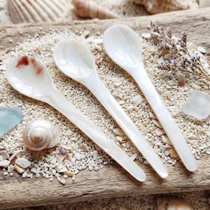 May include: Three white mother-of-pearl spoons with long handles are arranged on a wooden surface covered in sand and seashells. The spoons are pearlescent and have a delicate, elegant appearance. A seashell and sea glass are also visible.