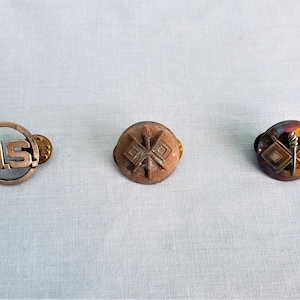 May include: Three vintage military pins. The first pin is silver and circular with the letters "US" inside. The second pin is brown and features a crossed flag design. The third pin is brown and features a crossed flag design with a spear in the center.