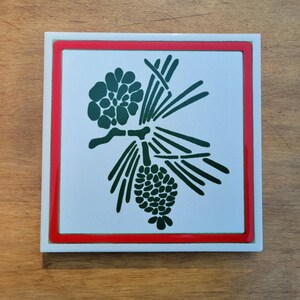 May include: Square ceramic tile with a white background, a red border, and a green pinecone and branch design. The tile is set against a wooden surface.