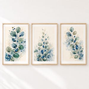 Könnte beinhalten: Ein Set aus drei gerahmten Wandbildern mit Aquarellillustrationen von Eukalyptuszweigen. Die Blätter sind in Salbeigrün, Petrol und Dunkelblau gehalten und mit kleinen goldenen Sprenkeln auf einem cremefarbenen Hintergrund akzentuiert.