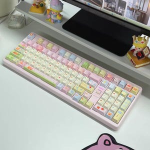Puede incluir: Un teclado mecánico rosa pastel con teclas coloridas. Las teclas presentan varios diseños, incluyendo frutas y patrones. También se ven un ratón y una alfombrilla rosas. Unas figuras decorativas están sobre un estante encima del teclado.