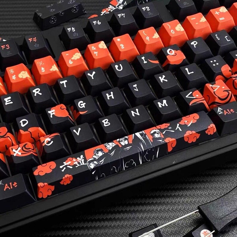 Pu&ograve; includere: Una tastiera meccanica nera con tasti personalizzati. I tasti presentano una combinazione di colori nero e rosso con disegni di ispirazione giapponese, tra cui nuvole e illustrazioni di personaggi. La tastiera ha un layout QWERTY standard.