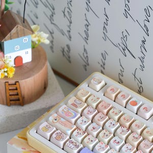 May include: A keyboard with custom keycaps featuring cat faces and other cute illustrations. The keycaps are in a light color palette with pastel shades. The keyboard is in a yellow tray, and a small wooden house is in the background.