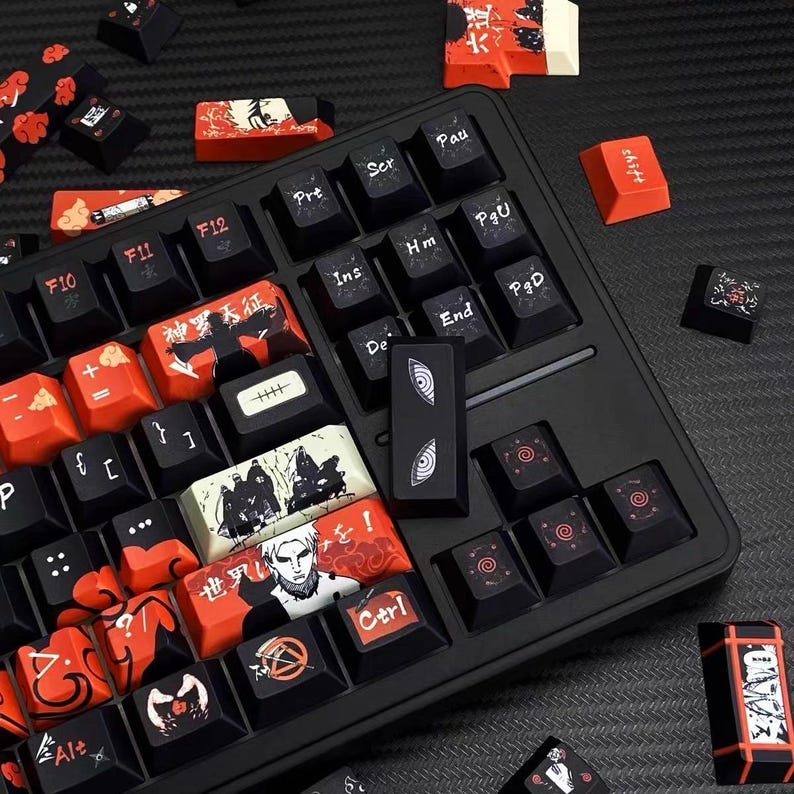 Pu&ograve; includere: Una tastiera nera con tasti personalizzati con design rossi e neri. Alcuni tasti presentano testo giapponese e illustrazioni in stile anime. I tasti sono sparsi intorno alla tastiera, mostrando i design unici.