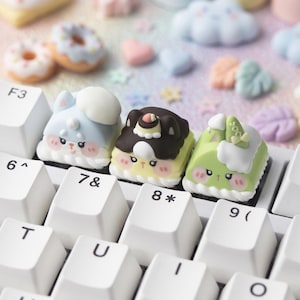 May include: Close-up of a keyboard with custom keycaps. The keycaps are designed as cute, colorful, cartoon-like characters, including a blue bunny, a brown bear, and a green creature. Other decorative elements are visible in the background.